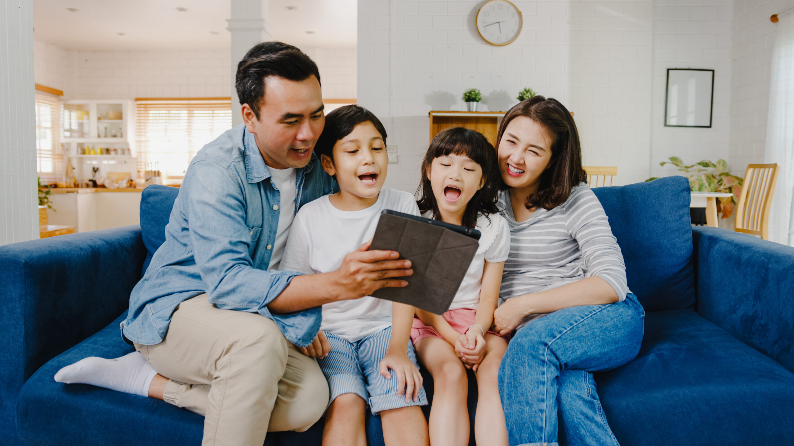 Happy Asian family (dad, mom, and two children) sitting on a blue sofa at home during school holidays, laughing and looking at a digital tablet together.
